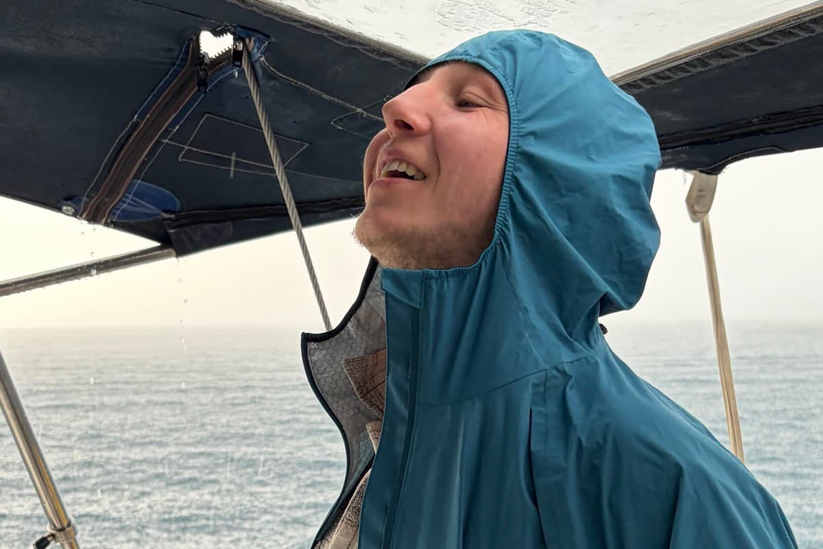 Ein Mann in Regenjacke lacht am Steuer im Regen, dahinter das Meer