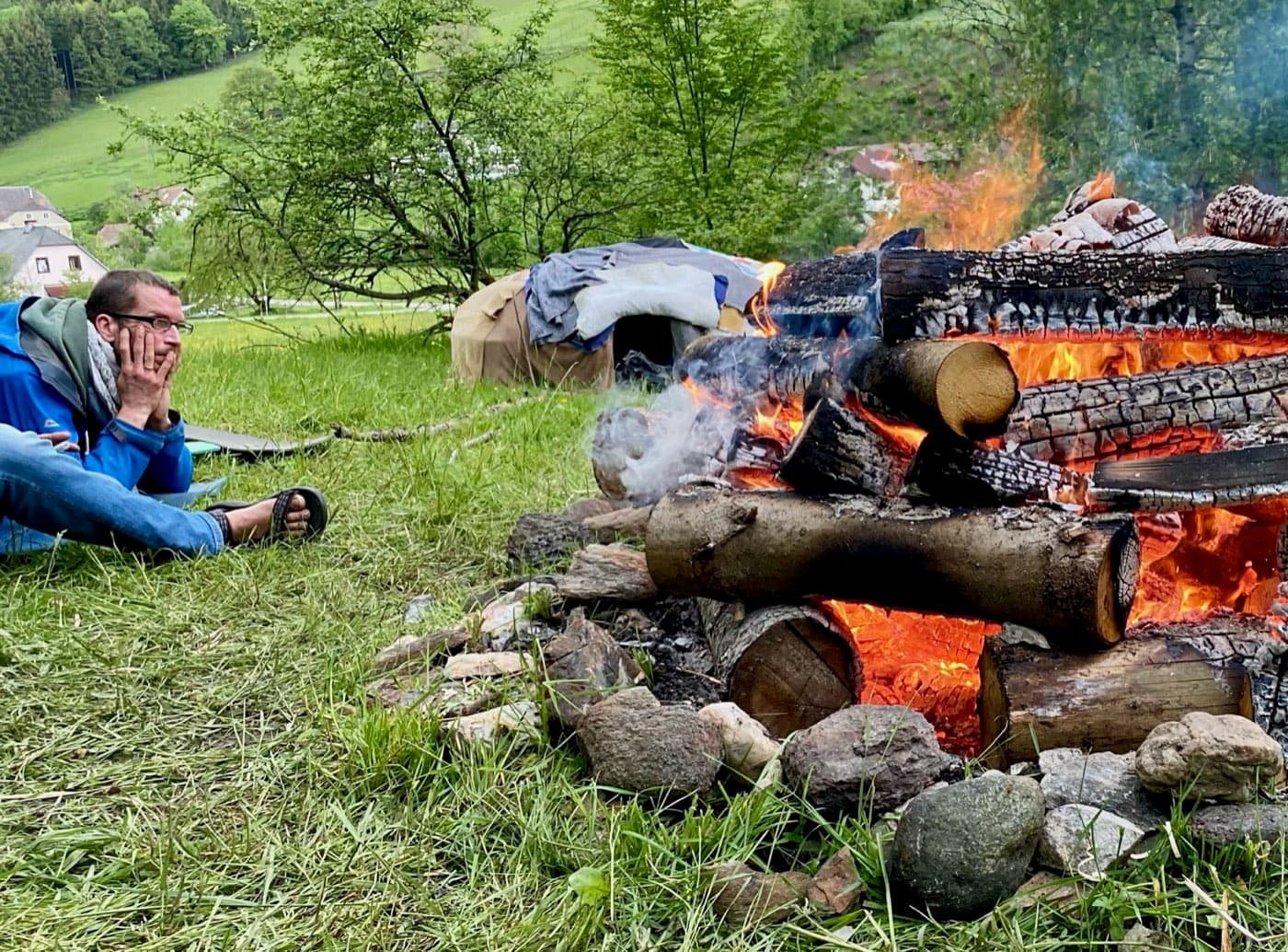 Ein Mann liegt im Gras und blickt in ein großes Feuer, dahinter die Schwitzhütte