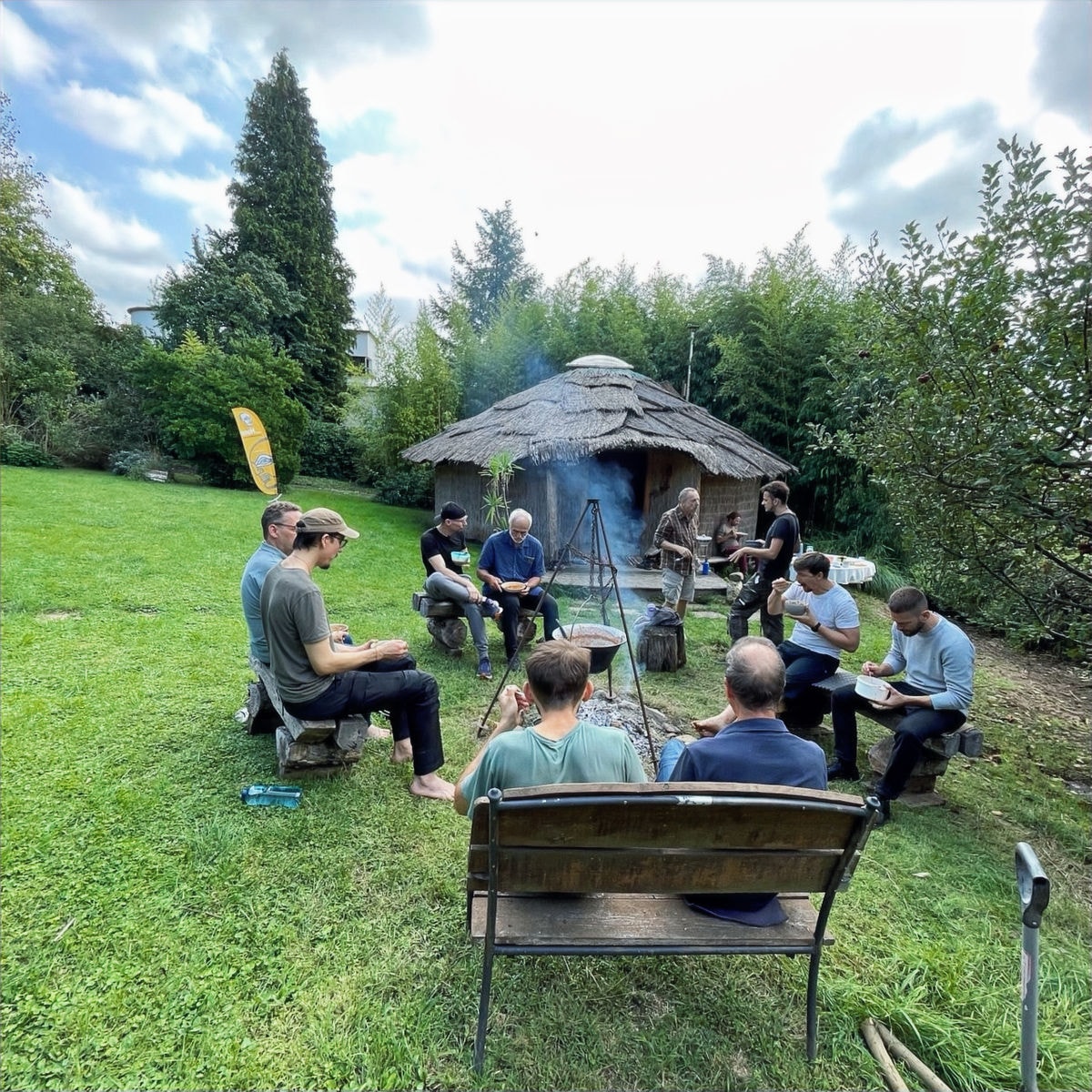 Männer sitzen entspannt im Kreis um ein Feuer vor einer strohgedeckten Hütte