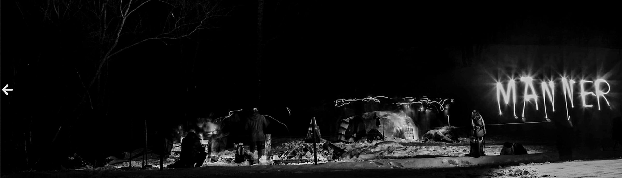 Nächtliches Winterlager im Schnee, Feuer und eine Schneehütte, im Hintergrund leuchtet das Wort „Männer" in Lichtschrift